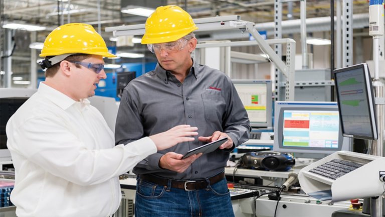 Dos hombres con cascos amarillos y anteojos de seguridad hablan entre ellos con una tableta en mano en un ambiente industrial